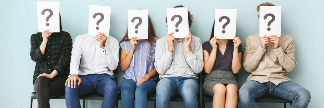 Young people hiding faces behind paper sheets with question marks while waiting for job interview indoors